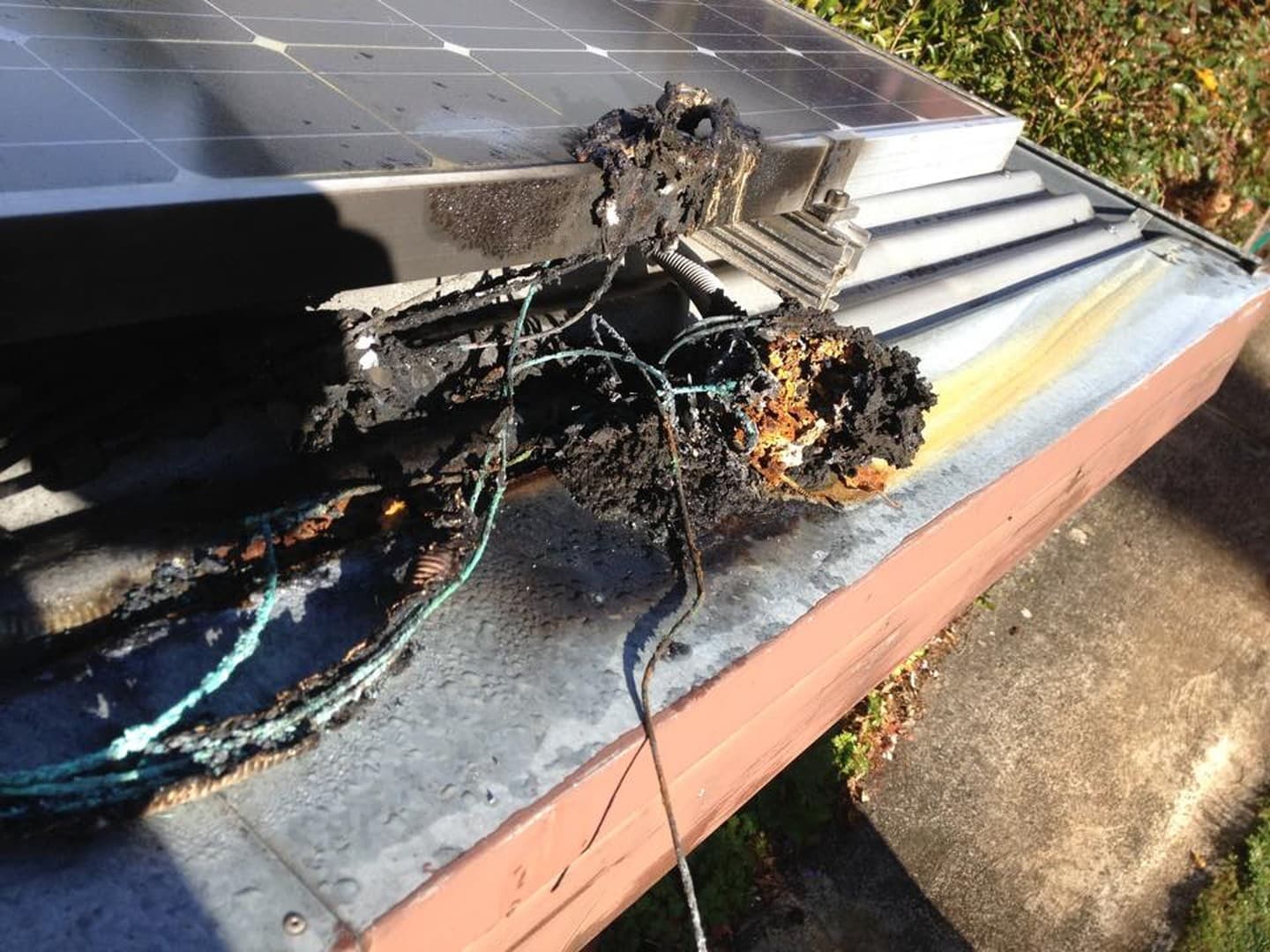A rooftop solar DC isolator burnt out at the edge of a solar panel, with charred cabling and exposed conductors on the roof edge