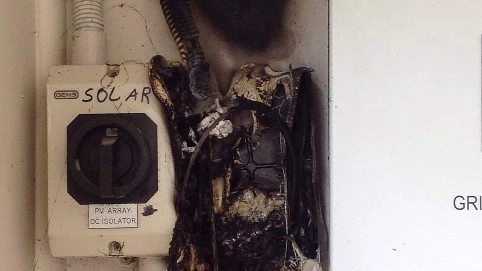 A burnt out solar DC isolator enclosure beside an intact isolator labelled PV Array DC Isolator, with soot up the wall