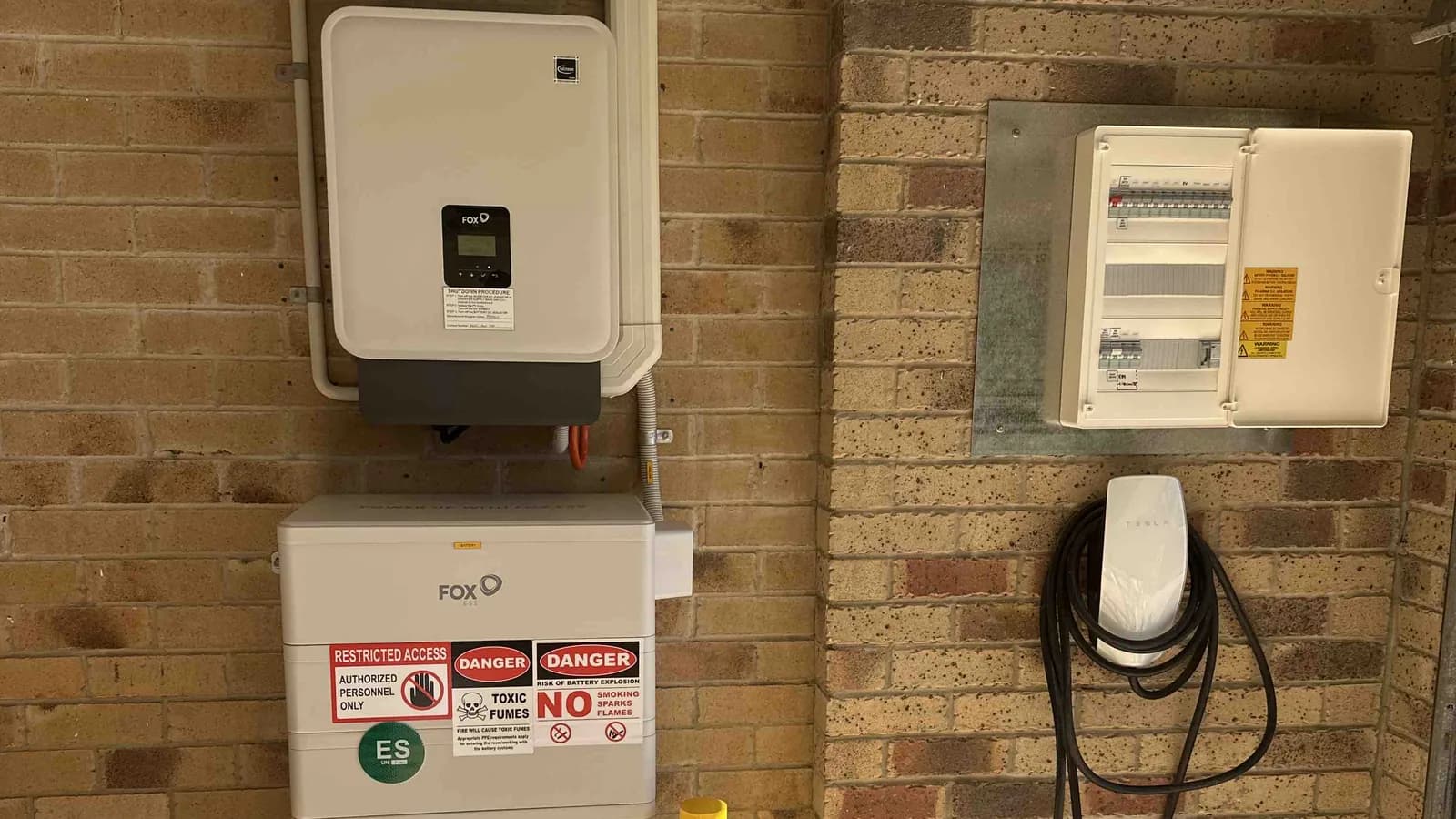 An EV wall charger installed on a brick wall beside a hybrid inverter, home battery and a sub board