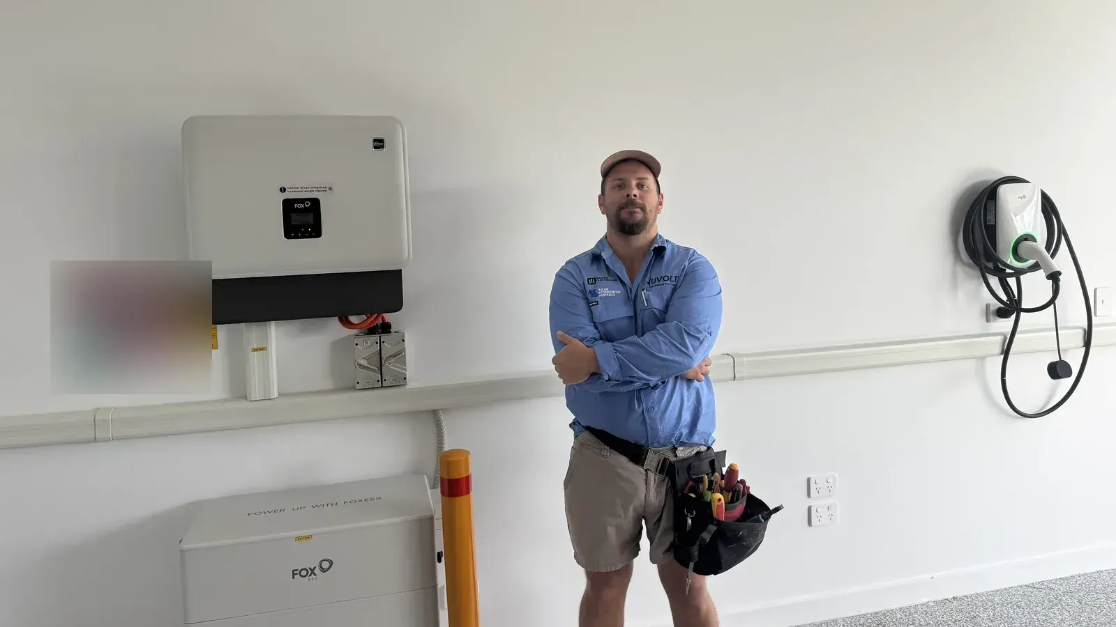 A Nuvolt electrician beside a Fox ESS hybrid inverter and CQ7 battery stack in a garage, with the cabling enclosed in ducting and a bollard protecting the battery