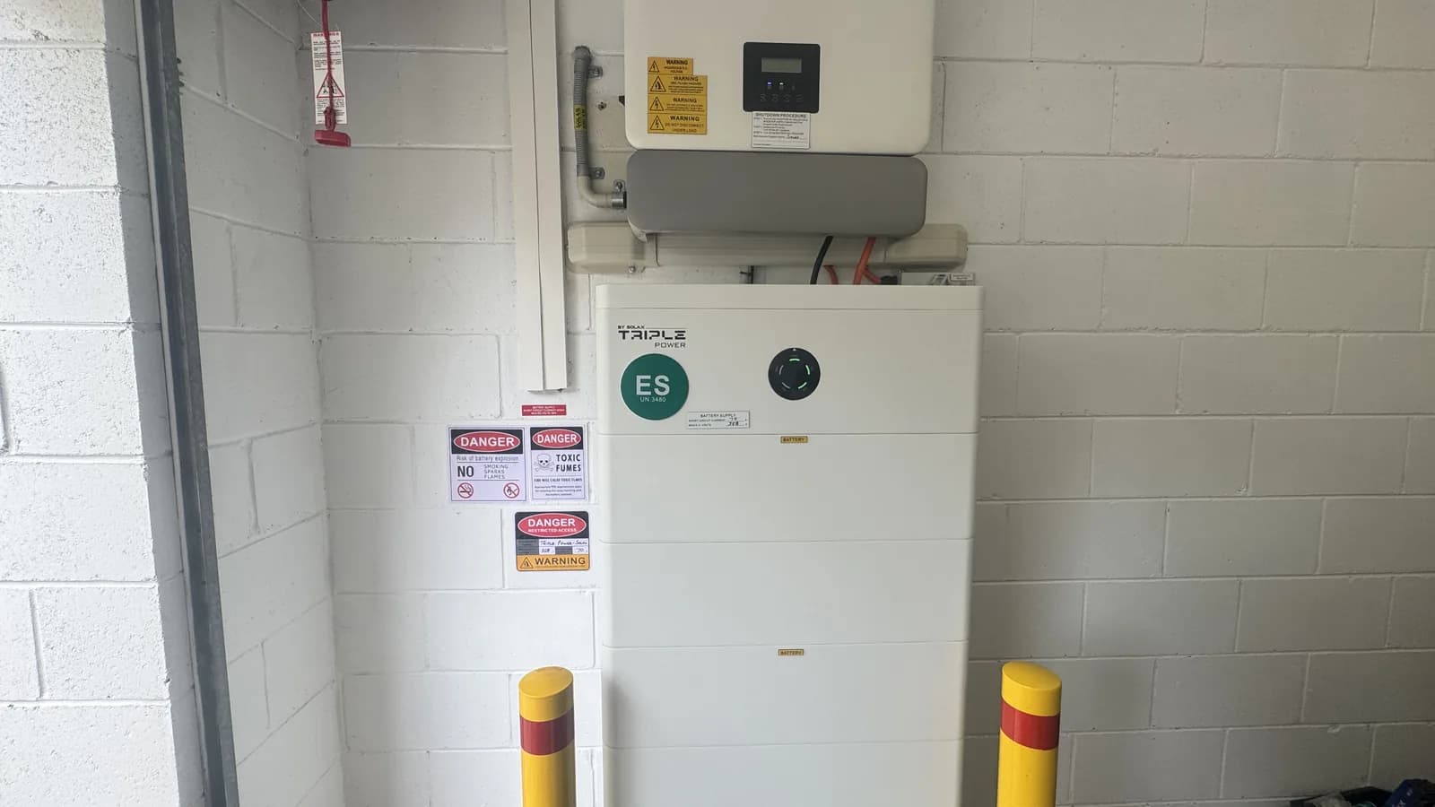 An installed home battery and hybrid inverter in a garage, with bollards and warning labels fitted