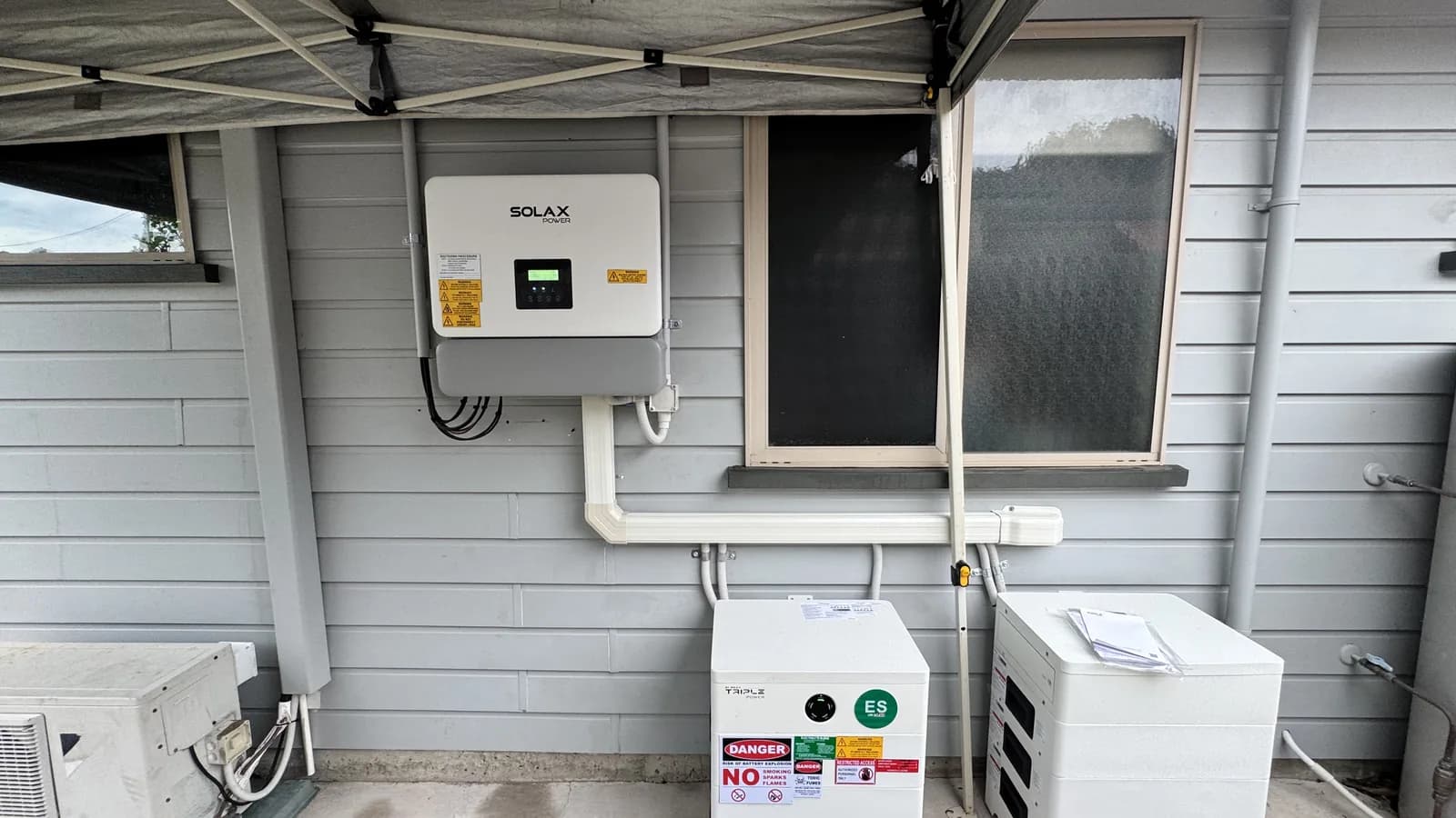 A SolaX hybrid inverter mounted beside a home battery on an external wall, wired back to the switchboard