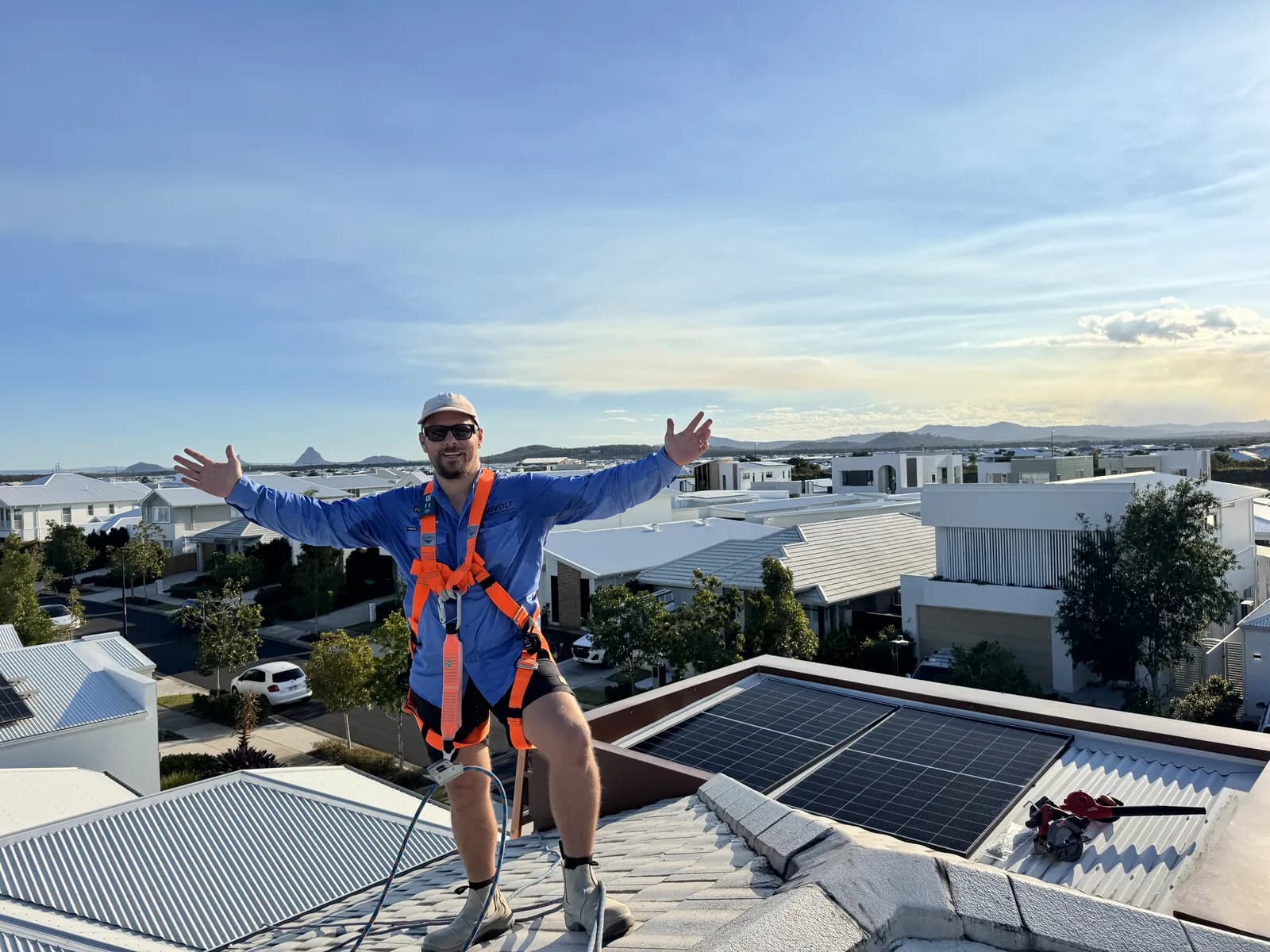 Nuvolt installer harnessed on a Sunshine Coast rooftop beside a finished solar array, with the Glass House Mountains behind