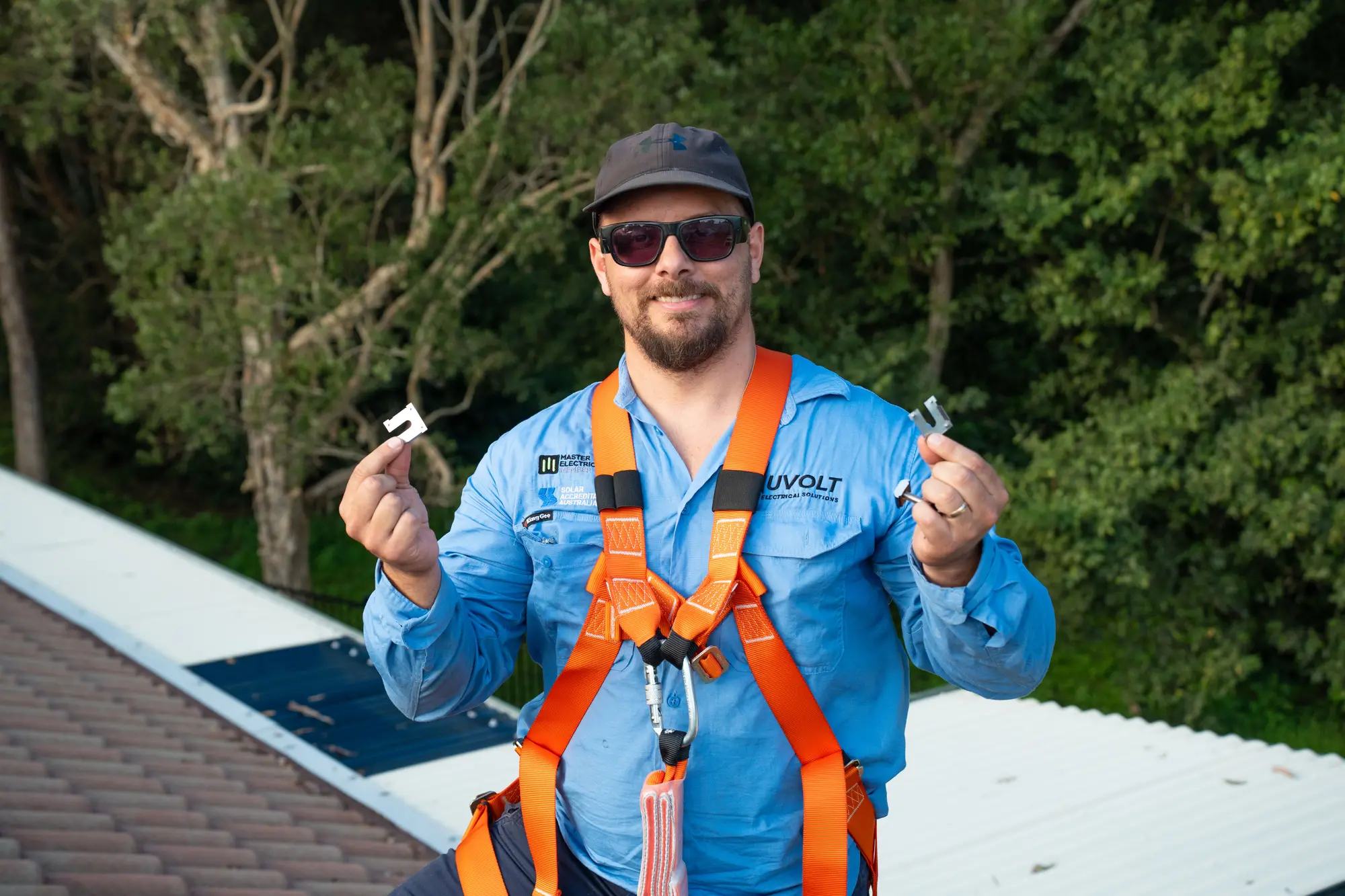 Dan from Nuvolt on a roof in a safety harness, smiling and holding up two solar panel clamps